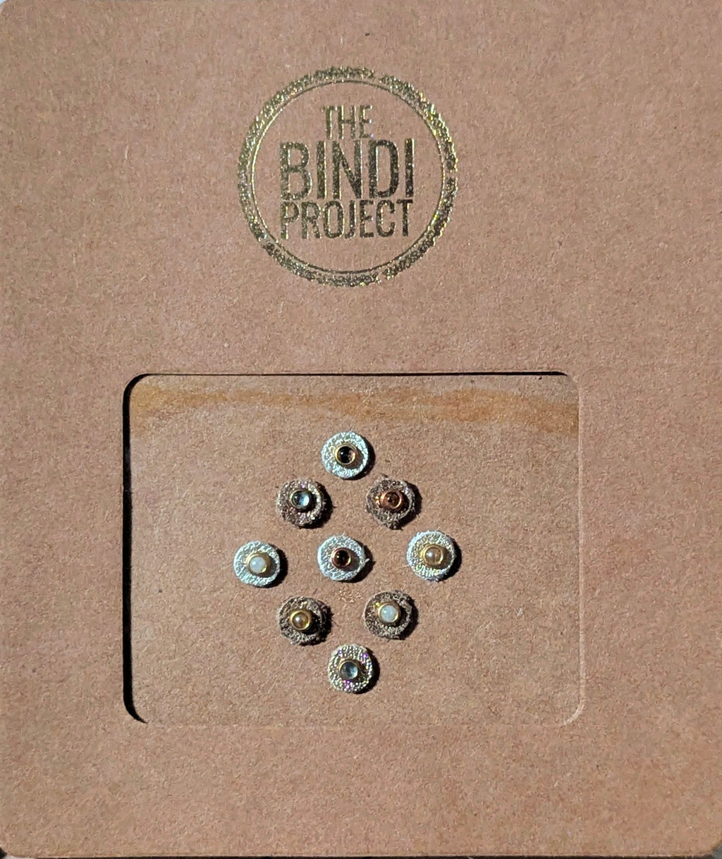 Nine small round artisanal bindis in neutral tones on textured brown card with green logo stamp.