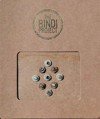 Nine small round artisanal bindis in neutral tones on textured brown card with green logo stamp.