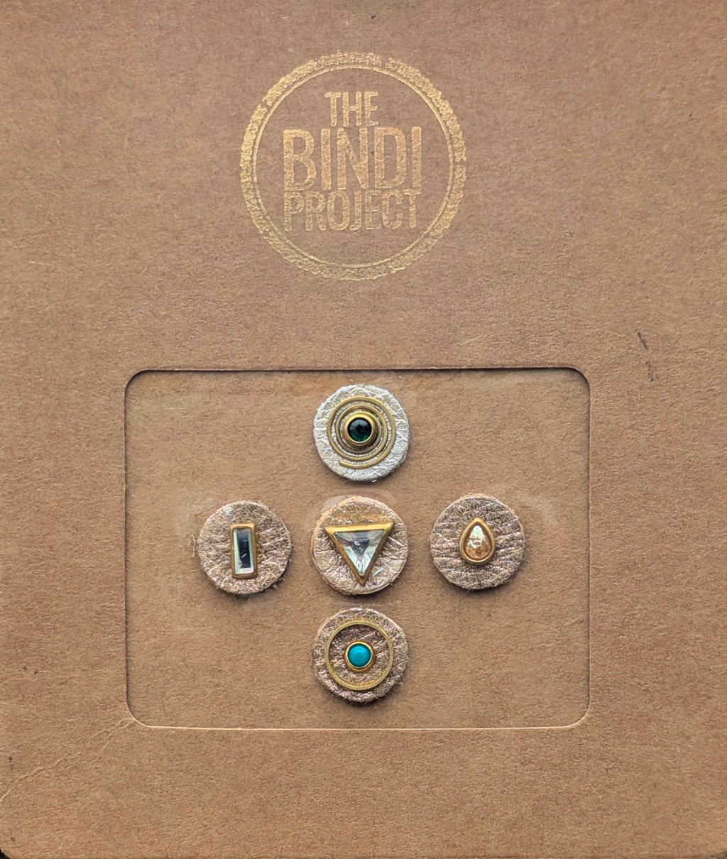 Five artisanal leather bindis in gold, turquoise, and black with geometric and teardrop designs on brown branded packaging.