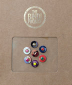 Seven colorful 10mm decorative bindis with textile and gemstone accents on brown packaging.